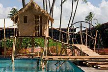 A grass and bamboo hut is sitting above a swimming pool due to bamboo stilts. A bridge made of wood is leading up from the right towards the hut. In the background is other buildings and blue, cloudy skies.