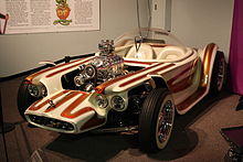 A white car with brown stripes, with open wheels and a clear bubble canopy over twin seats, and exposed, chromed engine with a blower. Beatnik Bandit II and a few of Roth's other cars are also on display in this museum.