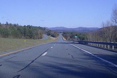 A four lane highway in the woods looking towards mountains on a sunny day.