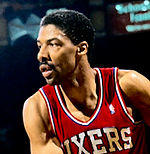A basketball player, wearing a red jersey with the word "SIXERS" on the front, is looking to his right side.