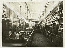 Photograph of a long narrow room taken from one end of the room. Open cabinets are hung on the walls for merchandise; there is a long counter that runs the length of the room. The room is illuminated by a long, glowing glass tube that rings the room and hangs a few feet down from the ceiling.