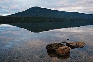 A calm lake reflects a partially clouded sky. A cluster of rocks protrude from the water at the shore near the viewer, while across the lake stands a wide, tree-covered mountain with a round peak.