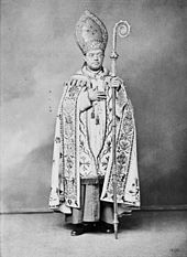 A bespectacled man holding a crosier in his left hand and wearing a mitre and liturgical vestments faces forward.