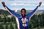 An African-American athlete wearing a blue running top and two gold medals hang from his neck.