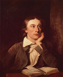 A man in his twenties looks to the right, resting his chin on his left hand with the elbow on a table in front of him. He has shoulder length brown hair and is wearing a brown jacket with a white shirt. A book is laying open on a table in front of him.