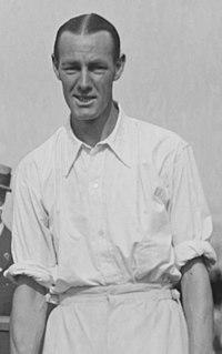 A black-haired man in a white shirt with sleeves rolled up looks into the camera, posing