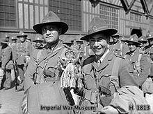 Two Maori officers wearing military uniforms smile at the camera, surrounded by other soldiers in front of a building