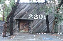Large, wooden building with a brown door (showing woodland animals play musical instruments) located in the bottom, centre left, and the large numbers "2200" painted in white above the door, centre-right. Asymmetrical trees with hanging foliage frame the building on all sides, while on the asphalt in the foreground, there are parking spaces and a disabled person sign.