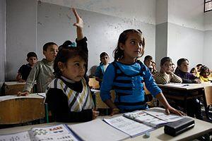 Children at desks in a classroom. One child raises her hand.