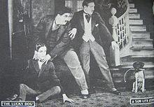 A promotional poster for Laurel and Hardy's film "Lucky Dog". Laurel is sitting on the left; Hardy is behind him with his hands around Laurel's neck. Both are looking at a small dog on the right.