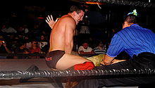 A dark-haired young man in a wrestling ring with black ropes. He is wearing black wrestling tights, with the name DiBiase written on them in red letters, and red kneepads with black wrestling boots. A referee wearing a blue shirt with thin black vertical stripes is in the corner.