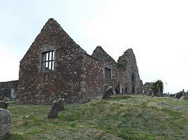 A photo of a ruined stone building with a gable and a mullioned window standing in a graveyard