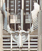 A stylized figure of a male human with outstretched arms and head tilted slightly forward, wearing a winged and crested helmet, mounted on the facade of a building