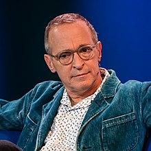 A bust photograph of a Caucasoid man in spectacles; he is wearing a white patterned shirt, blue jacket, and a jaw-mounted microphone. He is facing the camera, looking to its left.