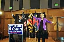 An older man and an older woman raise their hands together on a small stage in front of three flags.