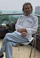 A middle-aged man with shoulder-long white hair, a white goatee, white blouse, and light-blue jeans. He is sitting cross-legged on a foldable chair on an outside balcony with potted plants placed in the background. Beyond the balcony, high-rise buildings and trees are visible.