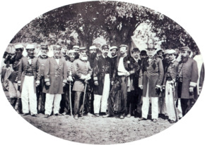A photograph showing a large group men, most of whom wear uniforms, standing beneath shade trees