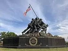 Color photo of the Marine Corps War Memorial, a bronze statue of six Marines raising a U.S. flag attached unto a Japanese pipe atop Mount Suribachi.