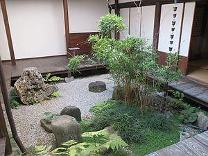 A garden courtyard with an en about 30 cm above the pale gravel, feature stones, a rain chain, and a planting of bamboo, ferns, grasses, and a creeping plant with small round leaves.