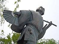 Statue of a woman with short hair, carrying a lady's fan and a samurai sword.