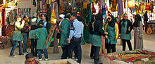 A group of young women, most in oversized green tunics and some wearing headscarves, in a bazaar. In front of them is a police officer in a blue uniform.