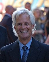 A man with long white hair and a black suit, with a tie, is standing and smiling.
