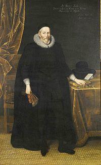 A tall elderly man with a beard, wearing long black robes and a large white ruff. He is standing with a fan in his right hand and with his left hand resting on books on a table.