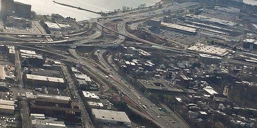 A series of highway ramps with multiple cars on them. A body of water is next to them and they are surrounded by buildings.