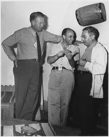 Three men talking. The one on the left is wearing a tie and leans against a wall. He stands head and shoulders above the other two. The one in the center is smiling, and wearing an open-necked shirt. The one on the right wears a shirt and lab coat. All three have photo ID passes.