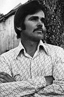 Photo portrait of a man with medium-length hair and a mustache crossing his arms and standing in front of a tree and a wooden shed