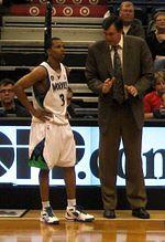 A man, wearing a brown suit, is talking to a basketball player on the side of a basketball court.