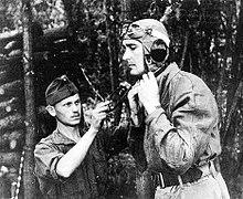 A man helps a taller young man to fix his flying helmet