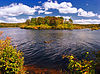 A sunny lake with an island and shore covered in autumnal foliage under a blue sky with some clouds