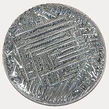 A shiny silver-white medallion with a striated surface, irregular around the outside, with a square spiral-like pattern in the middle.