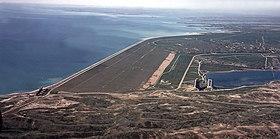 Aerial view of a large earthfill dam with its reservoir on the left and background, surrounded by brownish hills