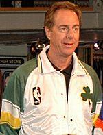 A man, wearing white and green jacket with the NBA logo and green shamrock on the front, is standing and looking to the front.