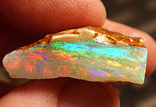 A small opal held side-on between someone's forefinger and thumb. A small sliver of brown rock is visible at the top, with a larger section of opal below it.