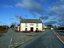 Front elevation of a white-painted rural pub with a central door, two lower and two upper windows edged with red, situated between two roads