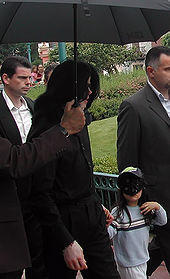 Jackson is wearing an overcoat and walking from left to right. His face is obscured by his hair. His son is wearing a mask and a baseball cap. Two men are with them; a third person is holding an umbrella over the Jacksons.