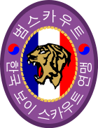 An oval badge of red with 범스카우트 (Tiger Scout) and 한국 보이 스카우트 연맹 inscribed in hangul; The center is an oval of yellow with the profile of a tiger head over a scroll inscribed with "preparation" and knot