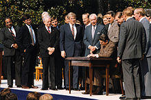 Rangel and about fourteen other men standing around a table, where President Ronald Reagan signs a bill