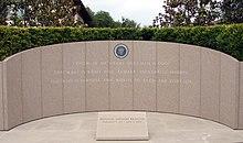A paneled, marble wall decorated with a seal and some inscribed words. A plaque with name and dates rests on the shiny marble floor.