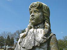 Grey stone statue of a 12 year old girl with shoulder-length hair.