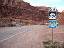 Signpost assembly against a red sandstone backdrop. The signs on the post read, "East, 128 (in a beehive shaped shield), and Scenic Byway".