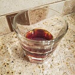 A small glass of clear brown liquid sits on a marble countertop.