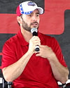 A man in his mid thirties speaking into a microphone. He is wearing a red and white baseball cap and a red t-shirt.
