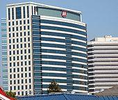 A white and blue building with a red logo on top stands behind and next to several short buildings.