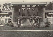 Low building with ivy draped terrace and three prominent vertical windows with tall trees in the background