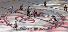 In the middle of a large red circle with the Toyota logo, lit partially by sunlight shining through the roof above, two hockey players from different teams are attempting to take control of the puck beneath them. The referee, in a black and white vertically striped shirt with an orange band around his upper right arm, stands off to their left. Around the edges of the circle are the two players' teammates, some just beginning to skate into the circle.