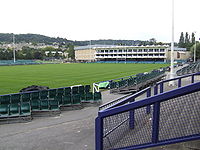 Area of mown grass with rugby posts and a white fronted pavilion building. In the foreground are terraces and seating with hills in the distance.